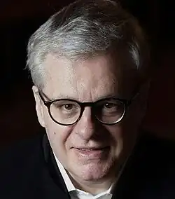 Dramatic, side-lit, colour publicity shot against a black background, of a half-smiling, clean-shaven, grey-haired, bespectacled man. His head is slightly bent downward, and he is looking upwards at the camera.