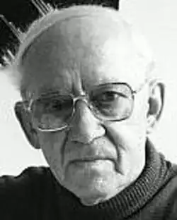 Black and white studio portrait of an elderly, unsmiling, clean-shaven, bespectacled man, with white hair and receding hairline.