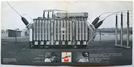 A black and white picture of an industrial generator in a field, with simple, one-story buildings in the background to the left. Underneath the generator is the album's cover, flanked by pictures of Hütter and Schneider, pictured separately, with additional album credits listed to the side of the photos.