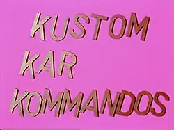 Bright pink screen with the words "KUSTOM KAR KOMMANDOS" in large, metallic-style capital letters arranged diagonally across the frame.