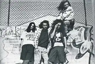 Four people, three of which are leaning against a wall and one of which is sitting on the wall. The wall has a lot of graffiti on it and there is a fence above the wall.
