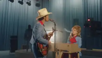 Bruno Mars and Lady Gaga in matching blue outfits on a TV studio set. Mars stands playing guitar while wearing a white cowboy hat. Gaga sits behind a piano with voluminous blonde hair, smoking.