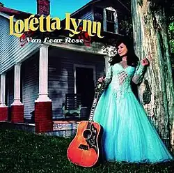 full-length photo of Loretta Lynn in light blue dress with guitar in front of house