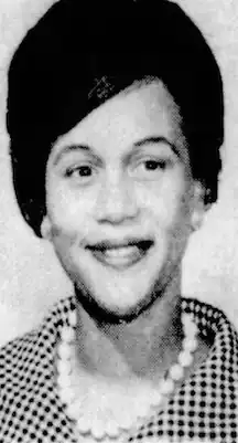 A smiling Black woman wearing a checked top and beads