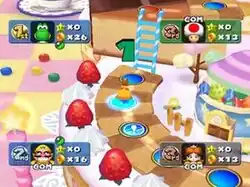 A horizontal rectangular video game screenshot that is a digital representation of a dessert-themed game board. A path made of checkered cookies sits on top of a giant white frosted cake with giant strawberries. A princess in an orange dress stands on a series of blue circles along the path. Character profiles with ordinal rankings and counters for stars and coins appear in each of the screen's corners.