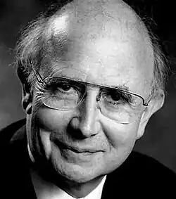 Black and white publicity shot of a clean-shaven, smiling, bespectacled man with short, grey, receding hair.