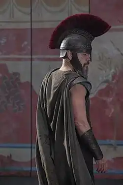 Picture of actor Matt Damon wearing a red-plumed Corinthian helmet, robed armor, and a gauntlet as Odysseus, standing in front of a wall with Greek murals.