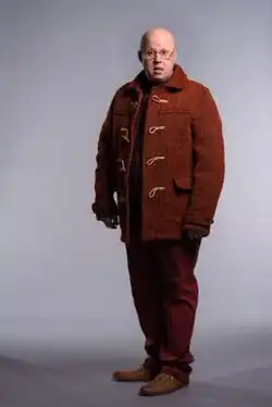 Nardole wears a red coat and stands against a grey background