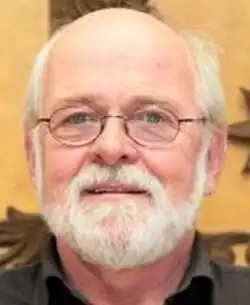 Colour Press shot of a half-smiling, elderly, bespectacled, balding man, with white hair and trimmed white beard and moustache.