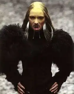 A woman from the waist up, wearing a black jacket with large furry sleeves, silver hoop earrings that touch her shoulders, and a mouthpiece that forces her lips open into a snarling expression