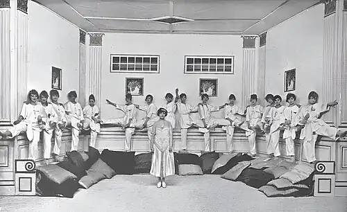 photo of young dark-haired white woman in white satin gown standing centre stage with chorus of 16 young women in white pyjamas spread out along the perimeter of the stage behind her
