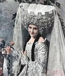 photo of young white woman in mediaeval costume, including long braids and heavy elaborate headdress, holding a few pink roses