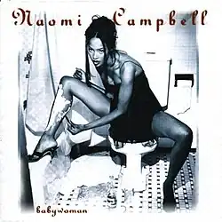 Naomi Campbell in a black dress seated on a toilet in a tiled bathroom, shaving her leg with shaving cream and a razor, with the title "babywoman" written at the bottom