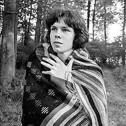 A black-and-white photograph of Nick Drake at age 18 or 19. He is standing in a field and has wrapped himself in patterned blanket.