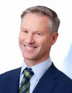 Middle-aged man with grey hair, wearing a navy blue jacket, white dress shirt, and necktie with a blue and gold-striped pattern
