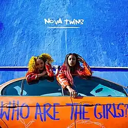 Two women leaning over the edge of a car with the words "WHO ARE THE GIRLS?" written on its side