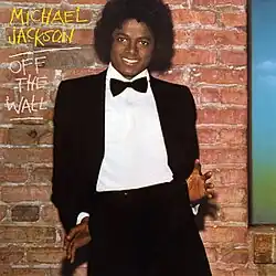 A smiling male (Michael Jackson) with a black afro, wearing a black tuxedo, white shirt, and a black bow tie. Both of his thumbs are hooked into his pants pockets with his palms and fingers facing forward and splayed out. The sides of his jacket are tucked behind his hands as he leans back slightly, giving a playful, casual touch to his formal look. Behind him there is a brown brick wall and to the side of his head are "MICHAEL JACKSON" in yellow chalk writing and "OFF THE WALL" in white chalk writing. "JACKSON" and "WALL" are separately underlined.
