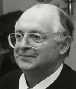 Black and white, three-quarter profile Press shot of a half-smiling, clean-shaven and bespectacled man with receding, greying hair.