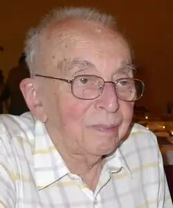 A balding elderly man in a collared shirt and glasses looking off camera
