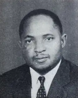 A black-and-white head-and-shoulders portrait of Aubrey Kachingwe. He is a man with short dark hair, looking directly at the camera with a neutral expression. He is wearing a dark suit jacket, a white collared shirt, and a patterned necktie. The background is a plain, textured grey.
