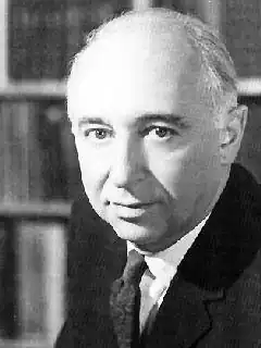 Black and white headshot of Simon. He looks into the camera with a neutral expression. He wears a dark suit and tie.