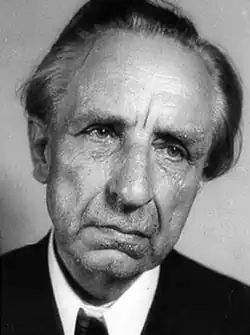 An elderly man in a suit with long hair pulled back