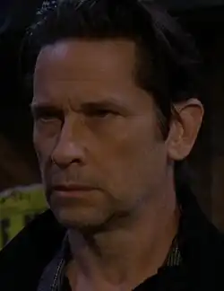 A dark-haired caucasian male in a top coat, scarf and silver chain.