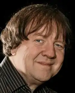 Colour, three-quarter profile publicity shot of a smiling, clean-shaven man with longish brown hair and fringe (bang)