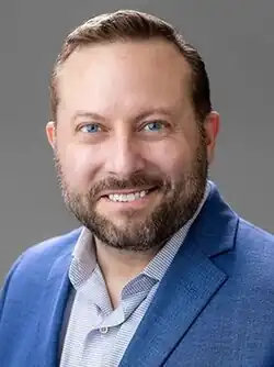 A portrait photograph of a white middle-aged man with short brown hair and blue eyes. He has a brown beard with some grey hairs in it. He is wearing a blue suit jacket over a white button-up shirt with blue stripes.