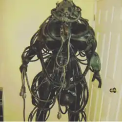 In front of a pale yellow wall stands a black, humanoid figure wrapped in several coiled wires