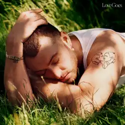 Sam Smith lying prone in grass with their head resting on their bent left arm and their right hand resting on their head. A tattoo of two men kissing is visible on Smith's shoulder.