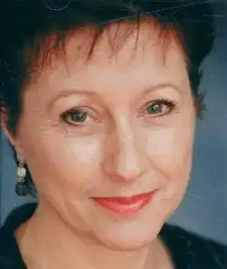 Colour, three-quarter profile, studio publicity headshot of a smiling woman with brown hair and coral pink lipstick.