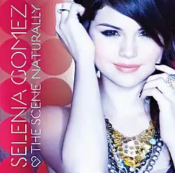 A portrait of a young woman in a circular, red background. She is tilting her hands to hold her black hair. The woman is wearing visibly pink lipstick, a gold-colour glitter-like necklace and a white sleevless top lined in black. On her right, horizontally vertical from her bottom right is the name 'Selena Gomez' in thin, white capital letters. Below it continues the name 'The Scene' and the title 'Naturally' in a reduced size of the same font.
