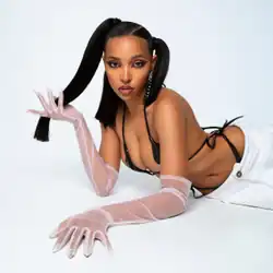 Tinashe lying on a white floor and looking at the camera, wearing a black string bikini top, white trousers, long sheer white gloves with exaggerated nails, and her hair styled in high pigtail ponytails
