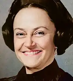 Hand-coloured, three-quarter profile, studio portrait of a smiling woman with backcombed brown hair, pink lipstick, and a drop earring.