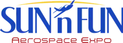 Logo depicts a small plane flying towards the viewer and facing the left. The background consists of two concentric circles (inner is yellow depicting the sun, outer is light blue to represent the sky). On the right is a palm tree and on the left (above the plane) is the name "SUN 'n FUN".