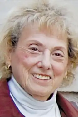 21st-century portrait photo of a smiling old woman with short, blonde hair, wearing a white turtleneck and red sweater