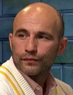 A White man wearing a pastel sweater vest over a pink button-down shirt.