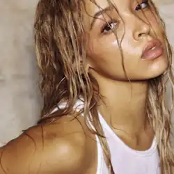 Tinashe looking up at the camera with wet hair, wearing a white ribbed tank top and thin hoop earrings against a light, textured background.
