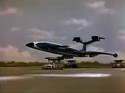 A blue, futuristic airliner rests on top of three orange support vehicles (one underneath the fuselage and two underneath the wings) as it speeds down an airport runway, the sky bright with clouds.