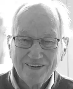 Black and white press headshot of a smiling, bespectacled, clean-shaven, elderly man with receding white hair.