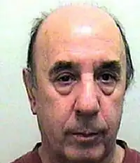 A photograph of William Goad following his arrest: he looks old with semi-balding hair line, bushy eyebrows, large bags under his eyes and some facial stubble. He has a white complexion with dark eyes and hair.
